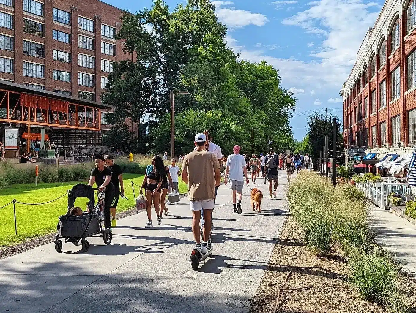 Pedestrians, a scooter rider, and a dog walker on the Atlanta Beltline urban trail, with Ponce City Market and converted brick warehouses lining the path.